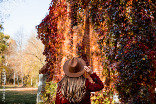 Young hipster millennials woman in a coat and hat in autumn nature. Model walks in the park and looks at golden autumn, colorful woods, fallen leaves, red loach wall. Autumn walk, people in fall.