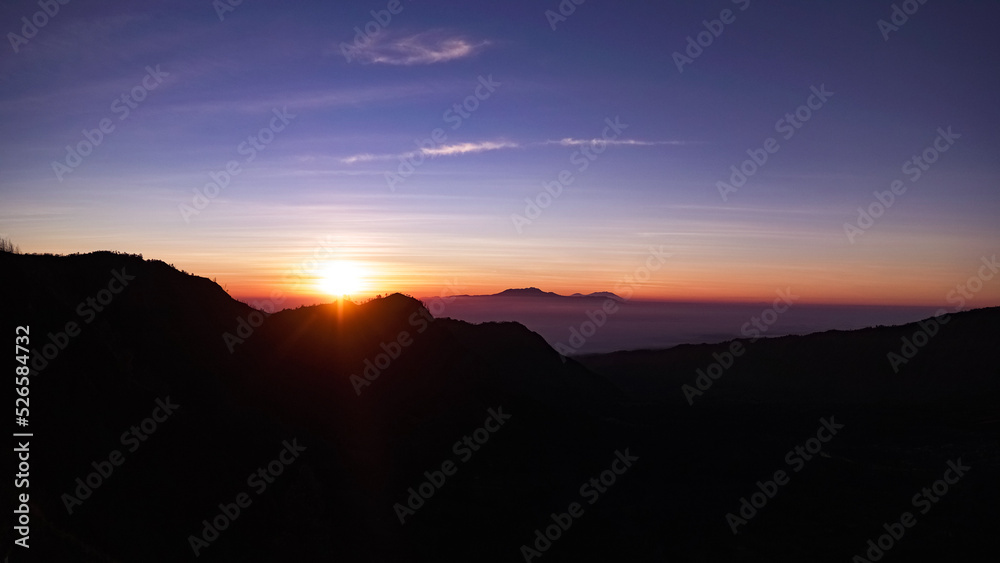 clear beautiful blue and orange sunrise with sun on the left over mountain silhouette with mist and clouds in the background on Bromo volcano landscape in Indonesia, Java island.