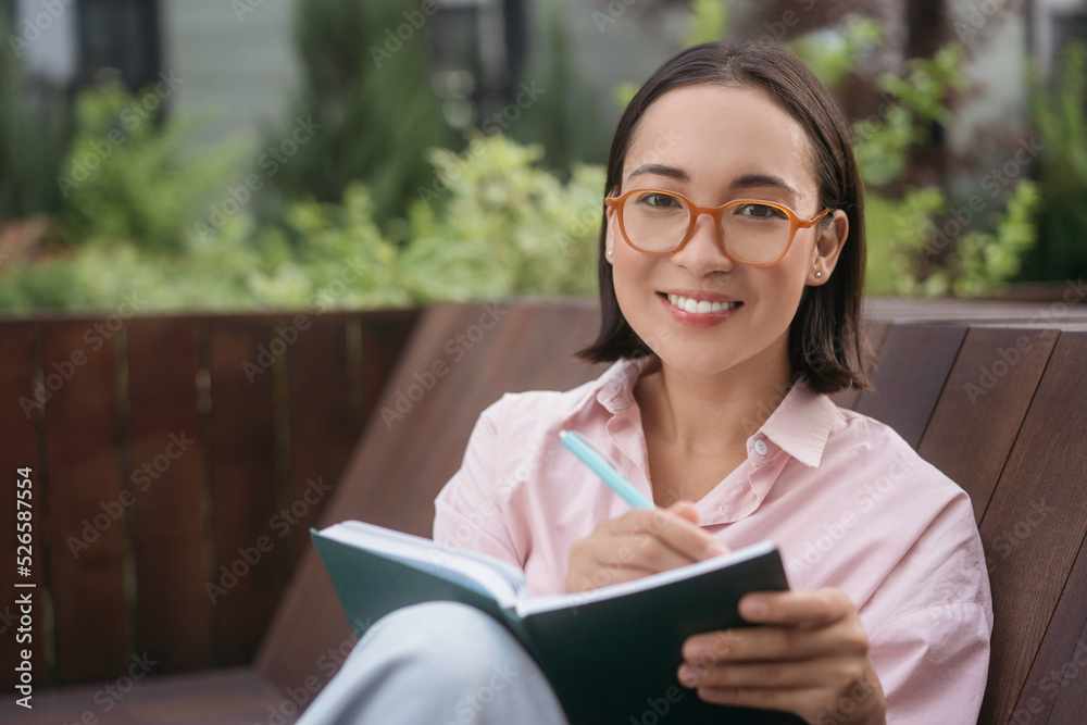 Portrait of young smiling asian writer wearing stylish eyeglasses looking at camera in park. Happy student studying, taking notes, reading book sitting in university campus. Education concept Portrait of young smiling asian writer wearing stylish eyeglasses looking at camera in park. Happy student studying, taking notes, reading book sitting in university campus. Education concept