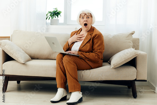 a shocked elderly woman with gray hair is sitting on a beige sofa in a brown suit working at a laptop and looking very emotionally amazed with surprise