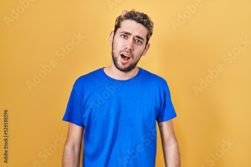 Hispanic man with beard standing over yellow background in shock face, looking skeptical and sarcastic, surprised with open mouth