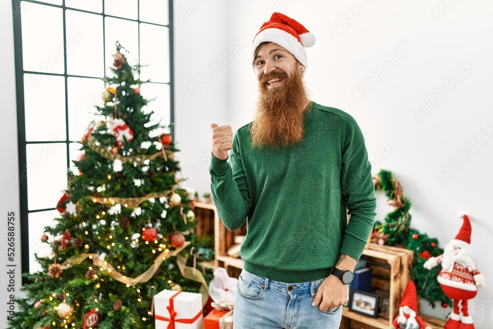 Redhead man with long beard wearing christmas hat by christmas tree smiling with happy face looking and pointing to the side with thumb up. Redhead man with long beard wearing christmas hat by christmas tree smiling with happy face looking and pointing to the side with thumb up.