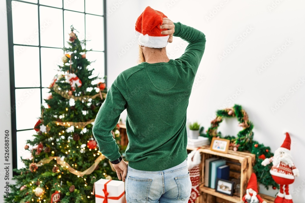 Redhead man with long beard wearing christmas hat by christmas tree backwards thinking about doubt with hand on head Redhead man with long beard wearing christmas hat by christmas tree backwards thinking about doubt with hand on head