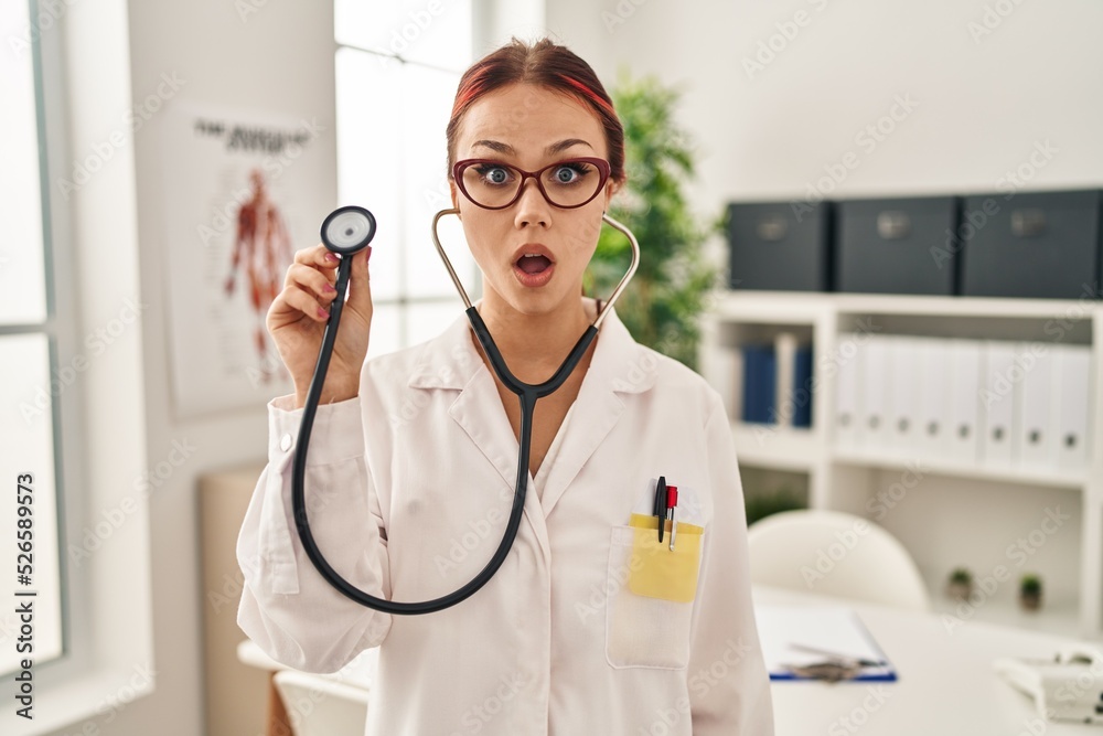 Young caucasian woman wearing doctor uniform using stethoscope scared and amazed with open mouth for surprise, disbelief face