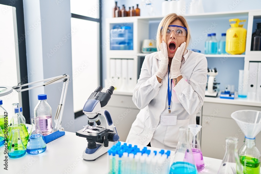 Middle age blonde woman working at scientist laboratory afraid and shocked, surprise and amazed expression with hands on face Middle age blonde woman working at scientist laboratory afraid and shocked, surprise and amazed expression with hands on face
