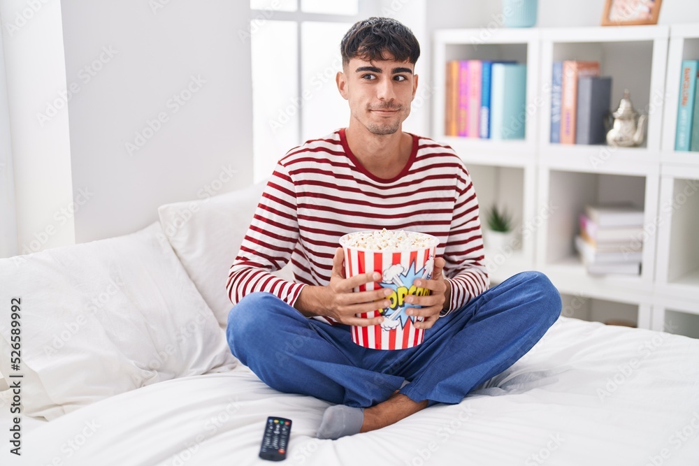 Young hispanic man eating popcorn sitting on the bed watching a movie smiling looking to the side and staring away thinking.