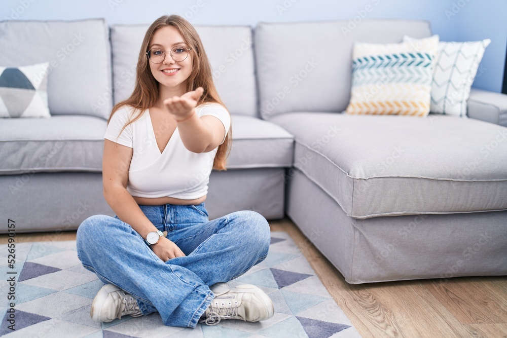 Young caucasian woman sitting on the floor at the living room smiling cheerful offering palm hand giving assistance and acceptance. Young caucasian woman sitting on the floor at the living room smiling cheerful offering palm hand giving assistance and acceptance.