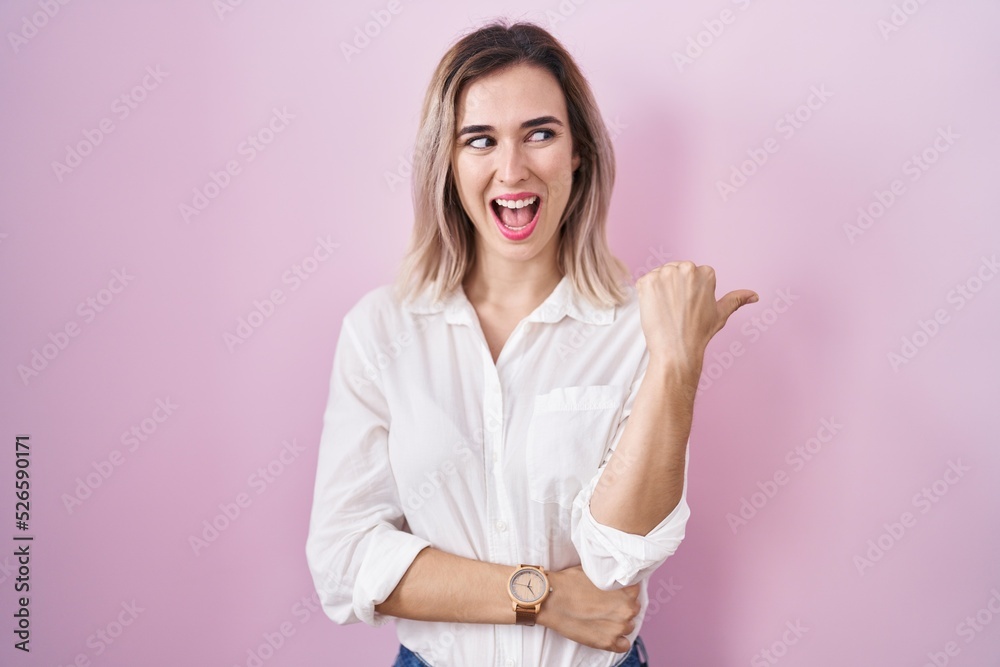 Young beautiful woman standing over pink background smiling with happy face looking and pointing to the side with thumb up.