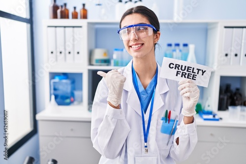 Young beautiful woman working on cruelty free laboratory pointing thumb up to the side smiling happy with open mouth