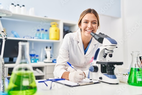 Young hispanic woman scientist using microscope write on document at laboratory