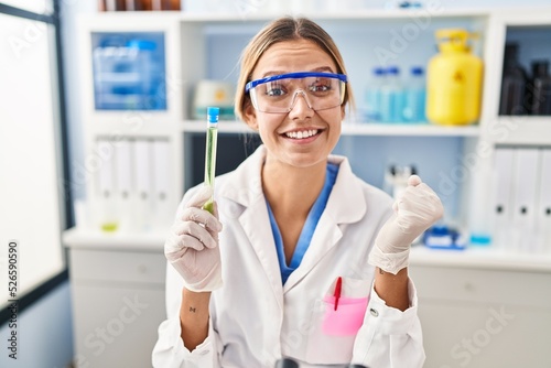 Young blonde woman working at scientist laboratory holding sample screaming proud, celebrating victory and success very excited with raised arm