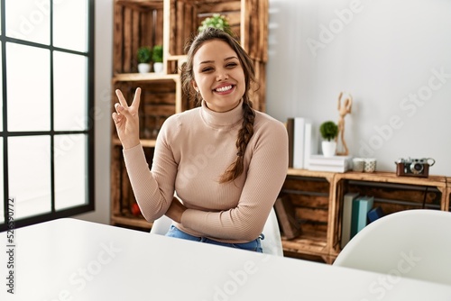 Young hispanic girl wearing casual clothes sitting on the table at home smiling with happy face winking at the camera doing victory sign. number two.