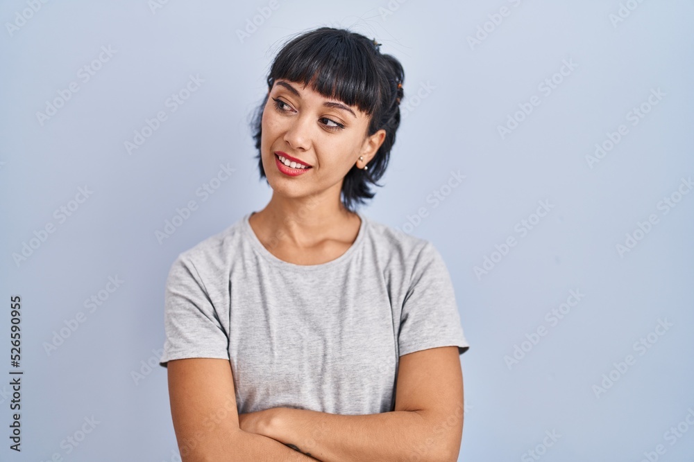 Young hispanic woman wearing casual t shirt over blue background looking away to side with smile on face, natural expression. laughing confident. Young hispanic woman wearing casual t shirt over blue background looking away to side with smile on face, natural expression. laughing confident.