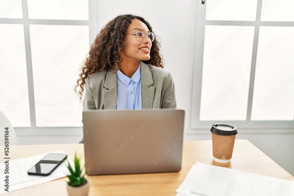 Beautiful hispanic business woman sitting on desk at office working with laptop looking away to side with smile on face, natural expression. laughing confident. Beautiful hispanic business woman sitting on desk at office working with laptop looking away to side with smile on face, natural expression. laughing confident.