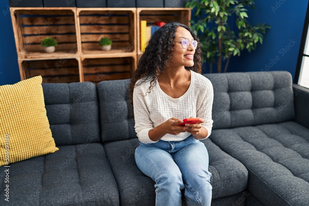 Obraz premium Young latin woman using smartphone sitting on sofa at home
