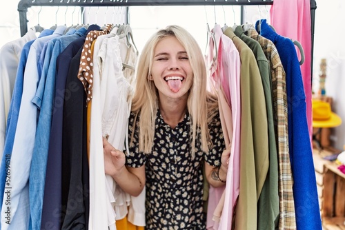 Young blonde woman searching clothes on clothing rack sticking tongue out happy with funny expression.