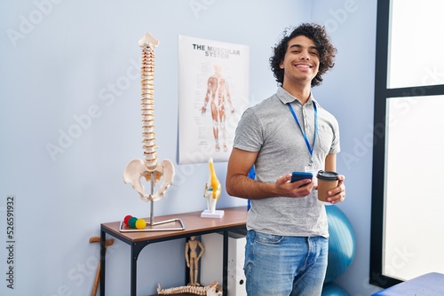Young hispanic man physiotherapist using smartphone and drinking coffee at rehab clinic