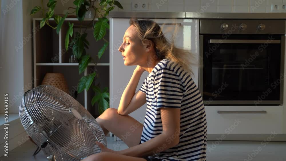 Exhausted young woman refreshing sit near big indoor ventilator blowing cooling fresh air at home on floor bonding. Tired female enjoying conditioner having fun indoors. Summer, heat weather concept