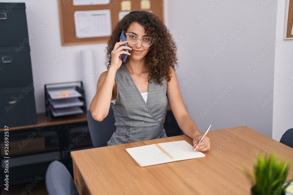 Young beautiful hispanic woman business worker talking on smartphone writing on notebook at office Young beautiful hispanic woman business worker talking on smartphone writing on notebook at office