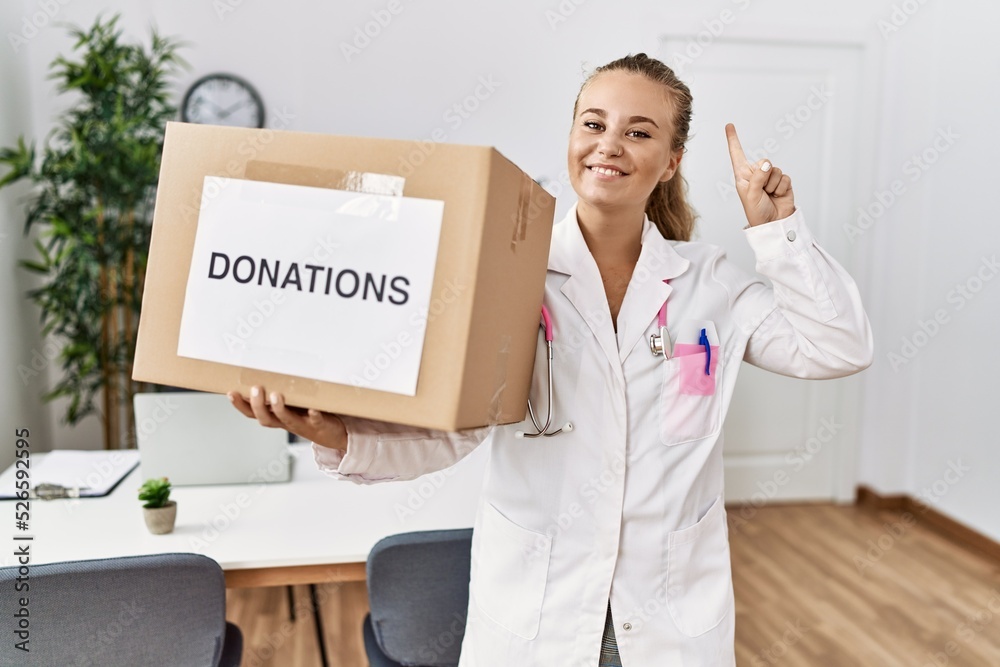 Young doctor woman at the clinic holding donations box smiling with an idea or question pointing finger with happy face, number one Young doctor woman at the clinic holding donations box smiling with an idea or question pointing finger with happy face, number one