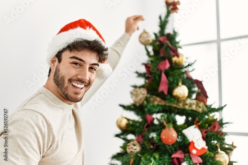 Young hispanic man smiling happy decorating christmas tree at home.