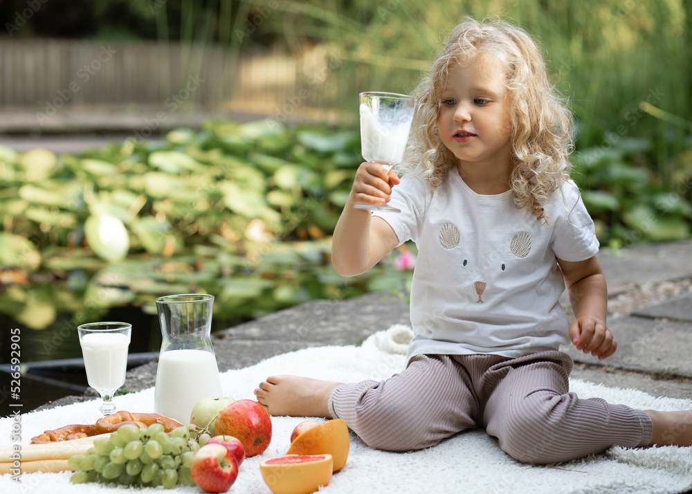 Portrait of amazing little girl with curly hair sitting on plaid on concrete pier near pond looking at glass of milk. Portrait of amazing little girl with curly hair sitting on plaid on concrete pier near pond looking at glass of milk.