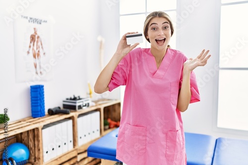 Young physiotherapist woman working at pain recovery clinic holding credit card celebrating victory with happy smile and winner expression with raised hands