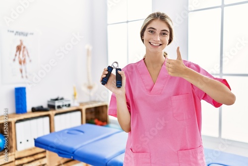 Young blonde woman working at pain recovery clinic holding hand strengthener smiling happy and positive  thumb up doing excellent and approval sign