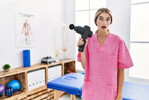 Young physiotherapist woman holding therapy massage gun at wellness center scared and amazed with open mouth for surprise  disbelief face