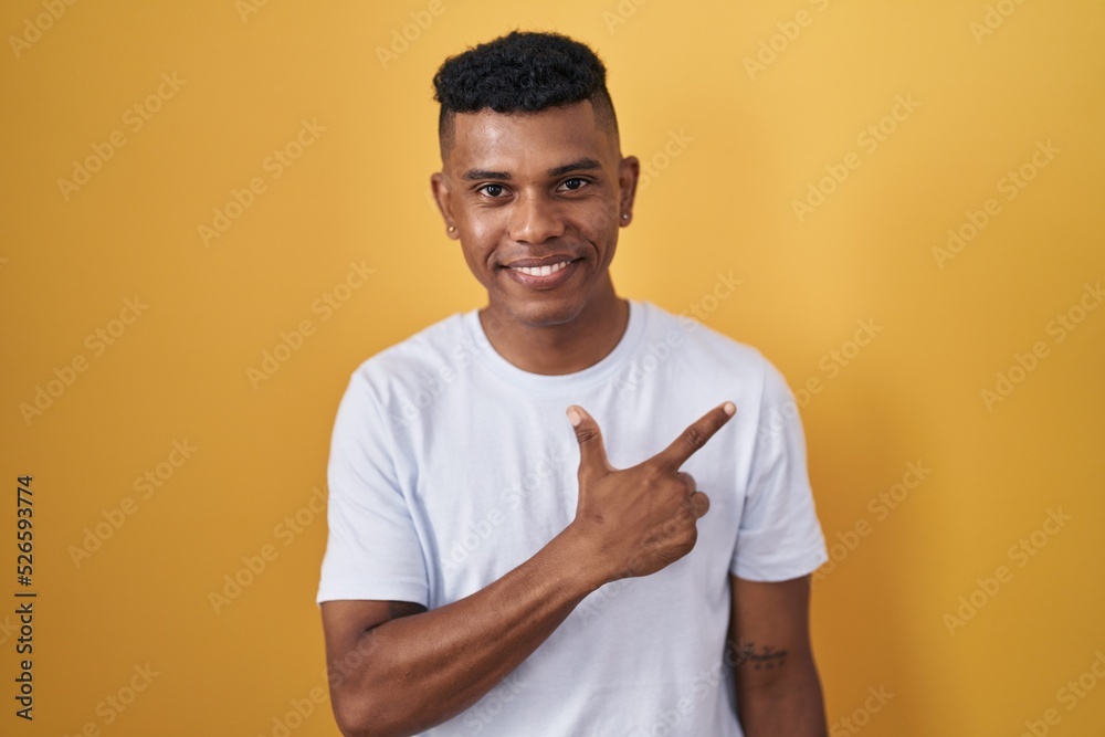 Young hispanic man standing over yellow background cheerful with a smile on face pointing with hand and finger up to the side with happy and natural expression