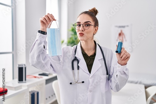 Young hispanic girl holding medical asthma inhaler at the clinic looking at the camera blowing a kiss being lovely and sexy. love expression.