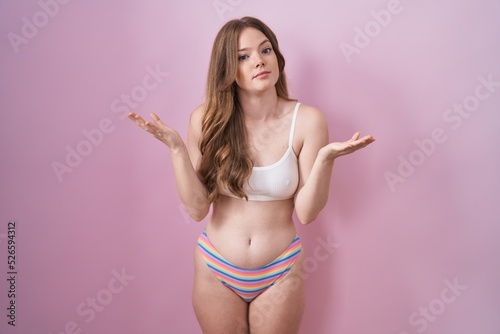 Caucasian woman wearing lingerie over pink background clueless and confused expression with arms and hands raised. doubt concept. © Krakenimages.com