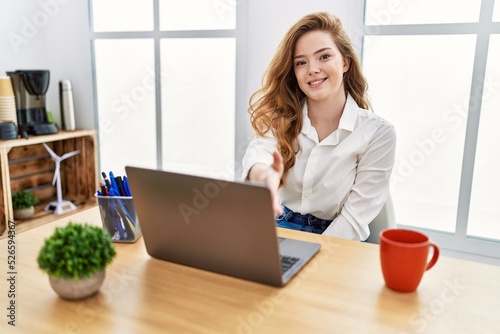 Young caucasian woman working at the office using computer laptop smiling friendly offering handshake as greeting and welcoming. successful business.