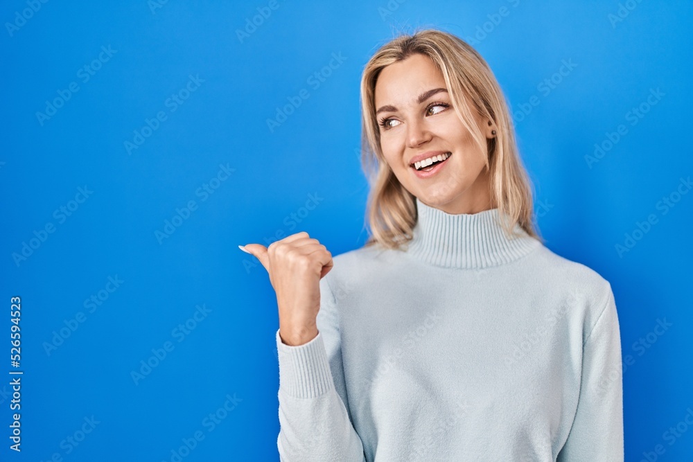 Young caucasian woman standing over blue background smiling with happy face looking and pointing to the side with thumb up. Young caucasian woman standing over blue background smiling with happy face looking and pointing to the side with thumb up.