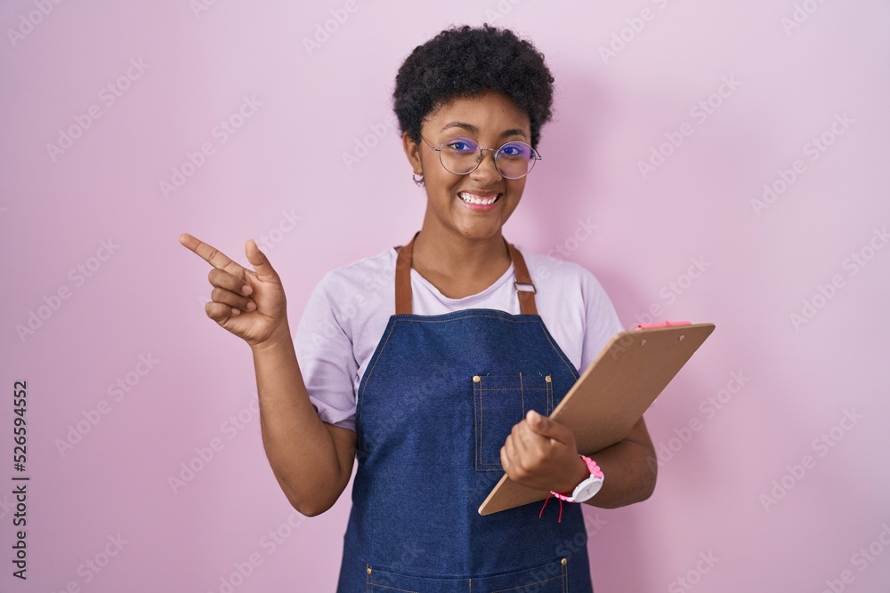 Young african american woman wearing professional waitress apron holding clipboard cheerful with a smile on face pointing with hand and finger up to the side with happy and natural expression