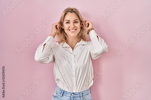 Young caucasian woman wearing casual white shirt over pink background smiling pulling ears with fingers, funny gesture. audition problem