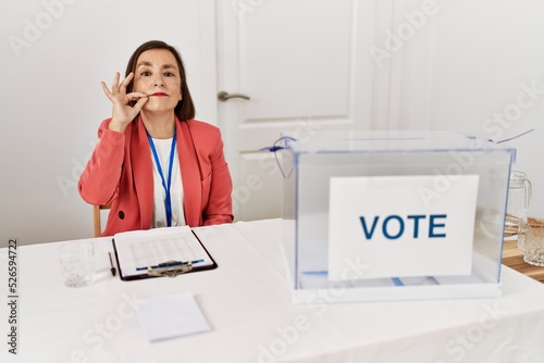 Beautiful middle age hispanic woman at political election sitting by ballot mouth and lips shut as zip with fingers. secret and silent, taboo talking photo