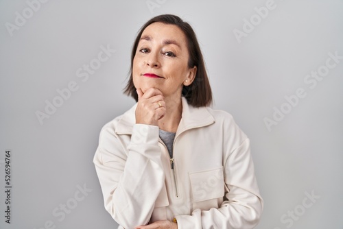Middle age hispanic woman standing over isolated background looking confident at the camera smiling with crossed arms and hand raised on chin. thinking positive.