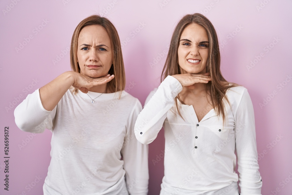 Middle age mother and young daughter standing over pink background cutting throat with hand as knife, threaten aggression with furious violence Middle age mother and young daughter standing over pink background cutting throat with hand as knife, threaten aggression with furious violence