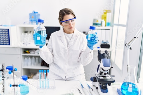 Young hispanic woman wearing scientist uniform holding liquid bottle at laboratory