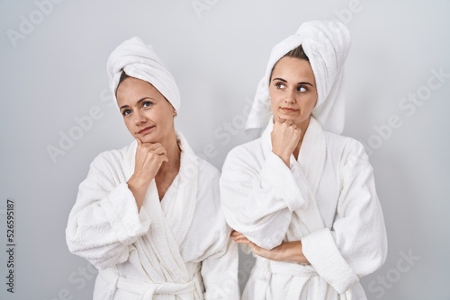 Middle age woman and daughter wearing white bathrobe and towel with hand on chin thinking about question, pensive expression. smiling with thoughtful face. doubt concept.