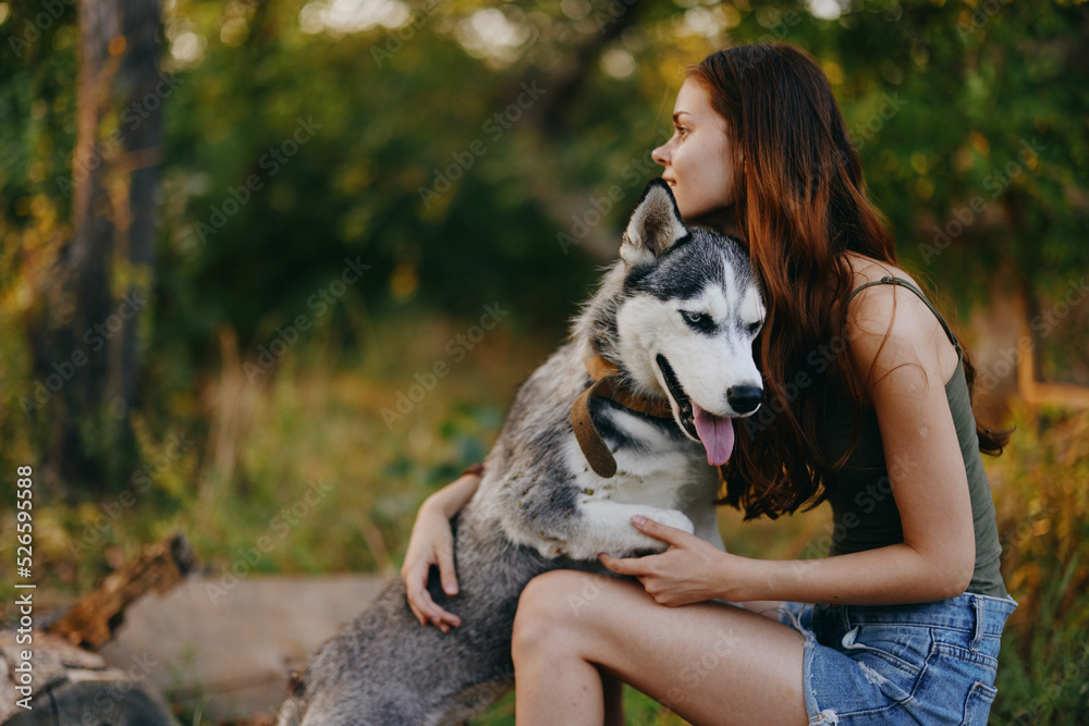 A young woman without makeup plays and smiles with her beloved husky dog ​​on a leash during a hike in the forest in autumn. Lifestyle together with a friend dog in nature