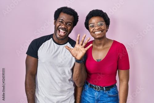 Young african american couple standing over pink background showing and pointing up with fingers number five while smiling confident and happy.