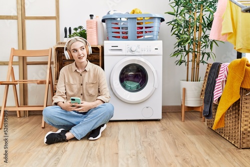 Young caucasian woman listening to music waiting for washing machine at laundry room