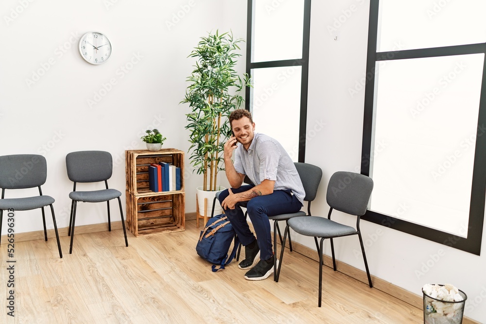 Young hispanic man talking on the smartphone sitting on chair at waiting room