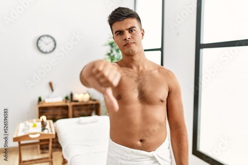 Young hispanic man standing shirtless at spa center looking unhappy and angry showing rejection and negative with thumbs down gesture. bad expression.