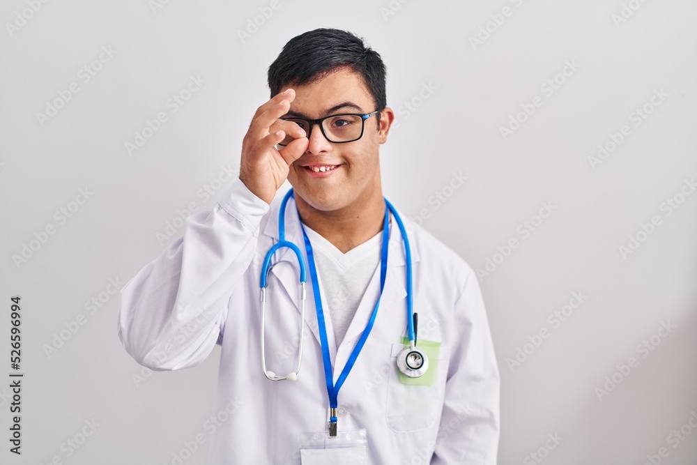 Young hispanic man with down syndrome wearing doctor uniform and stethoscope smiling happy doing ok sign with hand on eye looking through fingers Young hispanic man with down syndrome wearing doctor uniform and stethoscope smiling happy doing ok sign with hand on eye looking through fingers