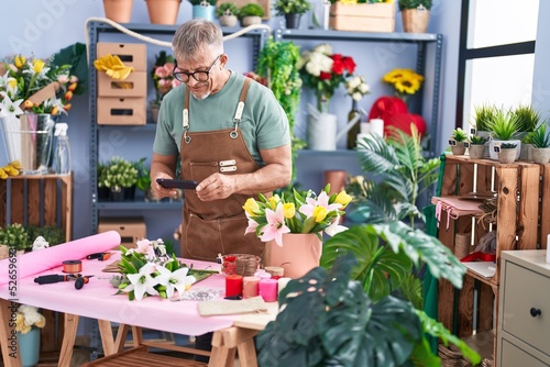 Middle age grey-haired man florist make photo to flowers by smartphone at flower shop