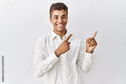 Young handsome hispanic man standing over isolated background smiling and looking at the camera pointing with two hands and fingers to the side.