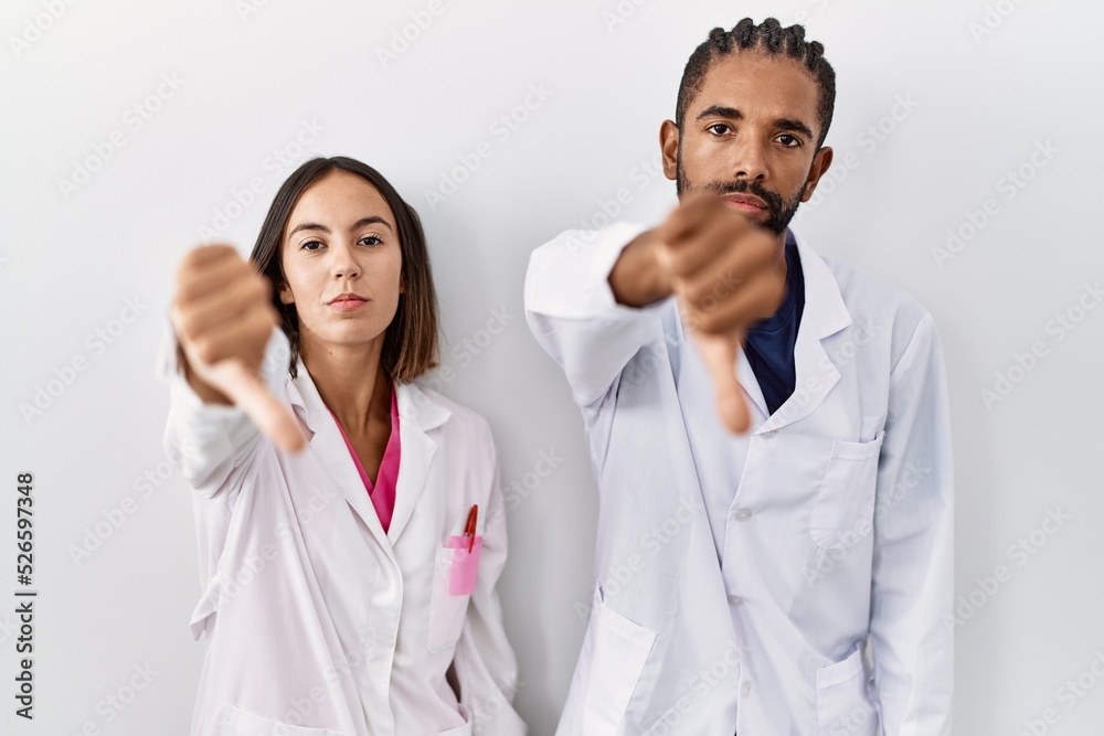 Young hispanic doctors standing over white background looking unhappy and angry showing rejection and negative with thumbs down gesture. bad expression. Young hispanic doctors standing over white background looking unhappy and angry showing rejection and negative with thumbs down gesture. bad expression.